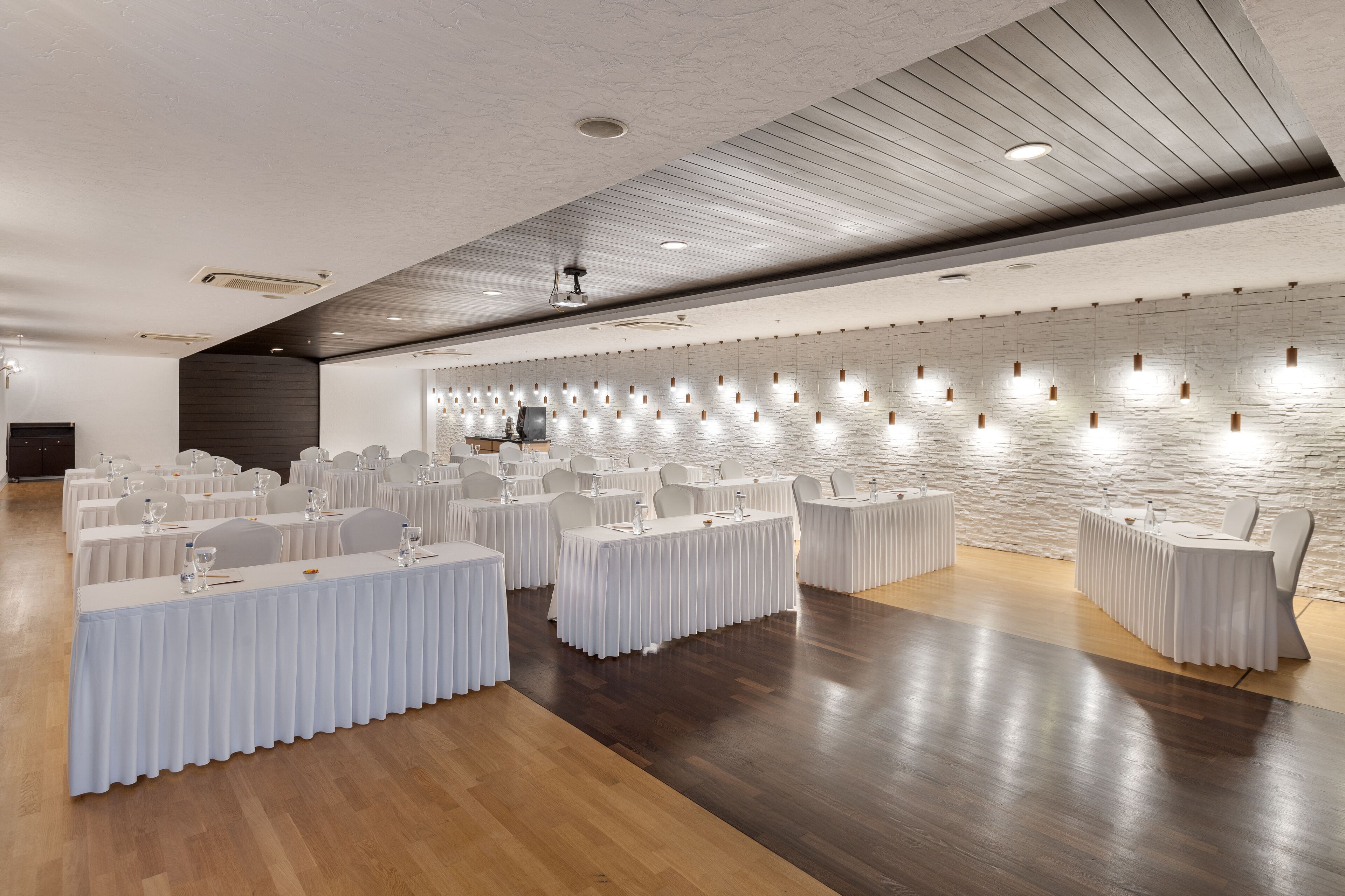Een ruime vergaderzaal met meerdere ronde tafels, witte tafelkleden en stoelen. De zaal heeft een houten vloer, een stenen muur met inbouwverlichting en een plafond met houten panelen en inbouwspots.