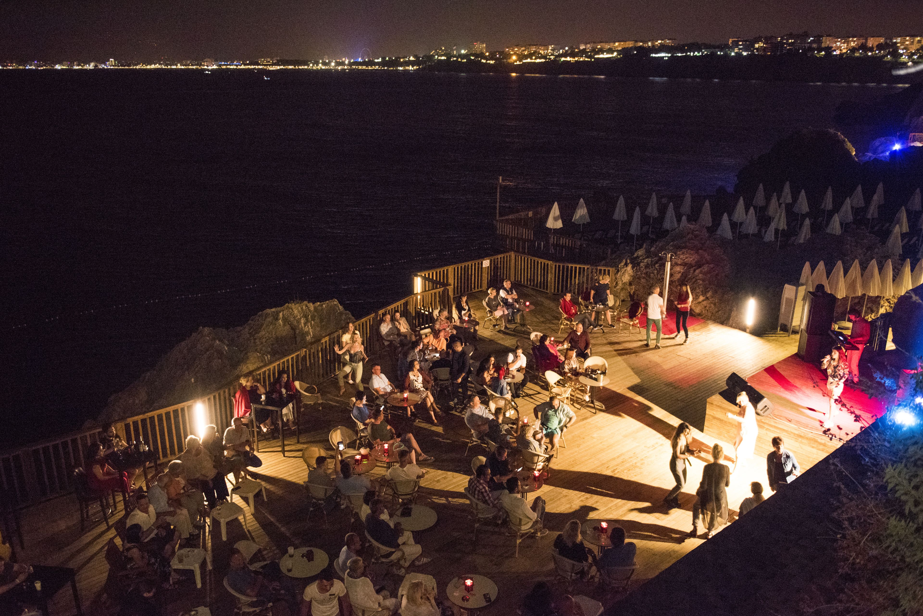 A deck with tables and chairs overlooking a body of water, with a stage and audience in the background.
