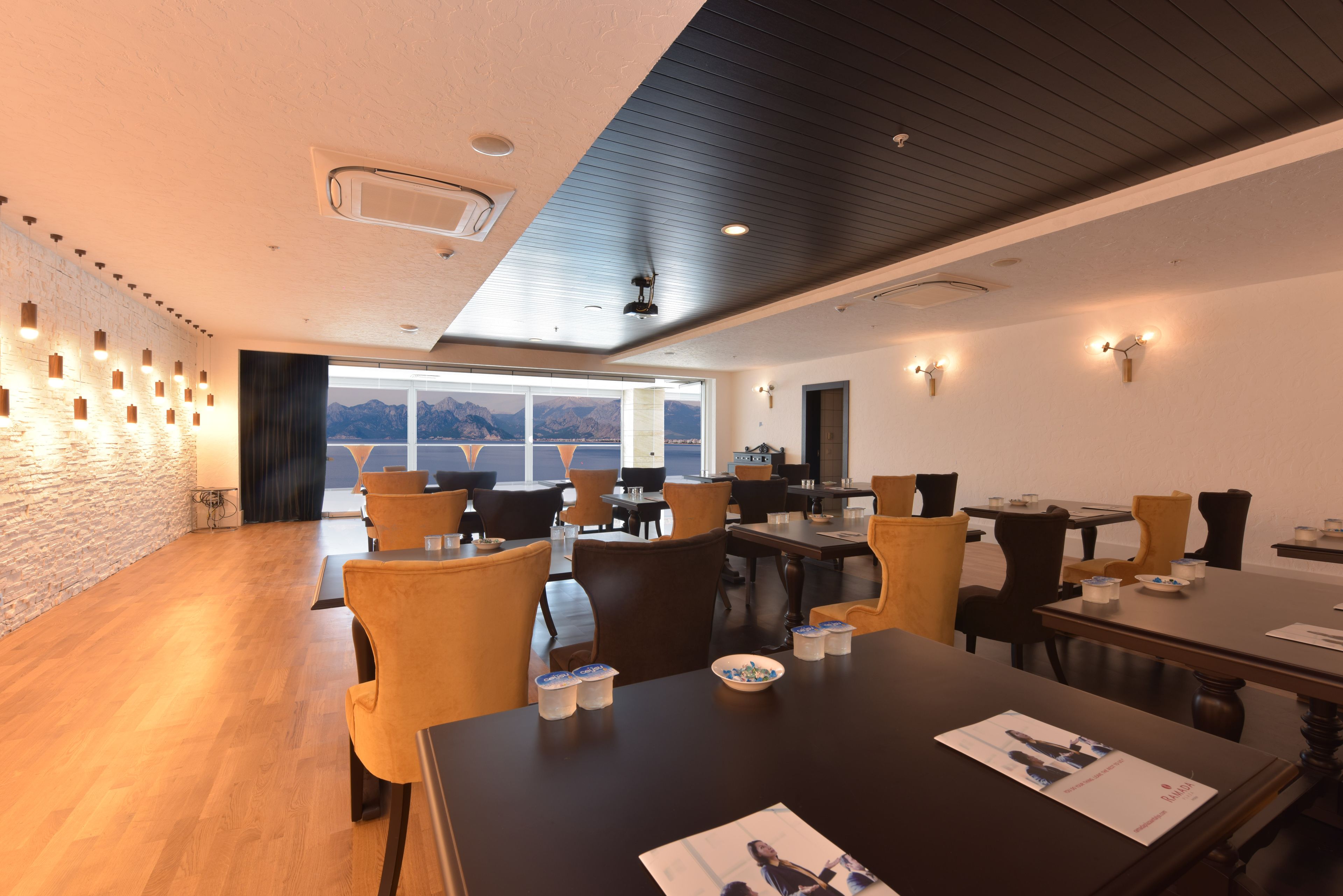 A modern dining area with wooden flooring, multiple tables and chairs, and a view of mountains through large windows.