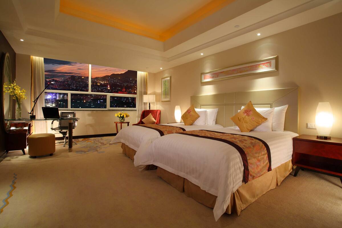 Ein Hotelzimmer mit zwei Betten, einem Schreibtisch, einem Stuhl, einer Lampe und Blick auf die nächtliche Stadt.