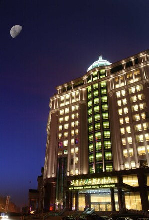Een hotel met een opvallende koepel en verlichte ramen, een bord met de tekst 'New Century Grand Hotel Changchun', en een zichtbare maan aan de hemel.