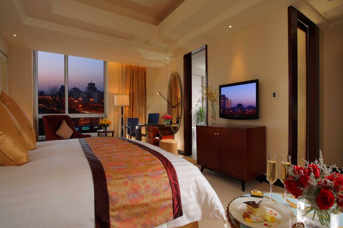 Ein Hotelzimmer mit einem großen Bett, einem Fernseher, einem Schreibtisch mit Stuhl, Blick auf die Stadtlandschaft und einem Esstisch mit Speisen und Getränken.