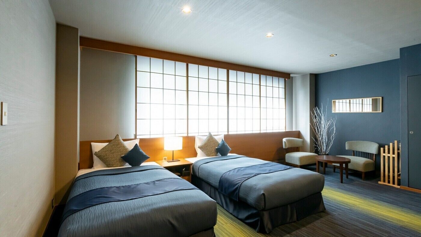 A hotel room with two beds, a sofa, a small table, and a window with blinds.