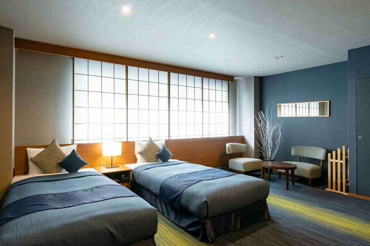 A hotel room with two beds, a sofa, a small table, and a window with blinds.