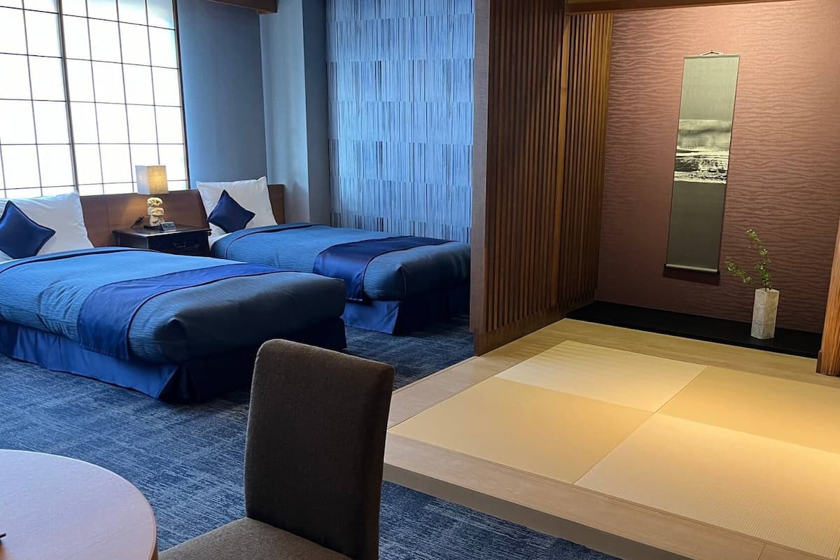 A hotel room with two beds, a small table, a chair, a TV, and a large window.