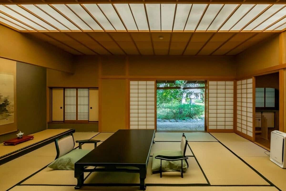 A traditional Japanese room with tatami flooring, a low table, a chair, sliding doors, and a view of greenery outside.