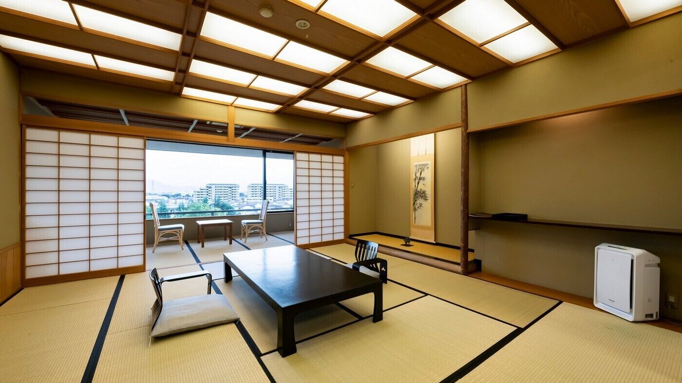 A traditional Japanese room with tatami flooring, a low table, chairs, a shoji screen, and a view of buildings.