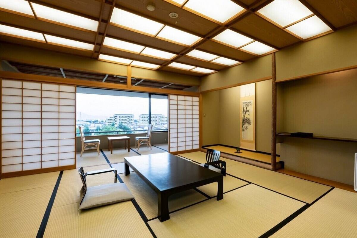 A traditional Japanese room with tatami flooring, a low table, chairs, a shoji screen, and a view of buildings.