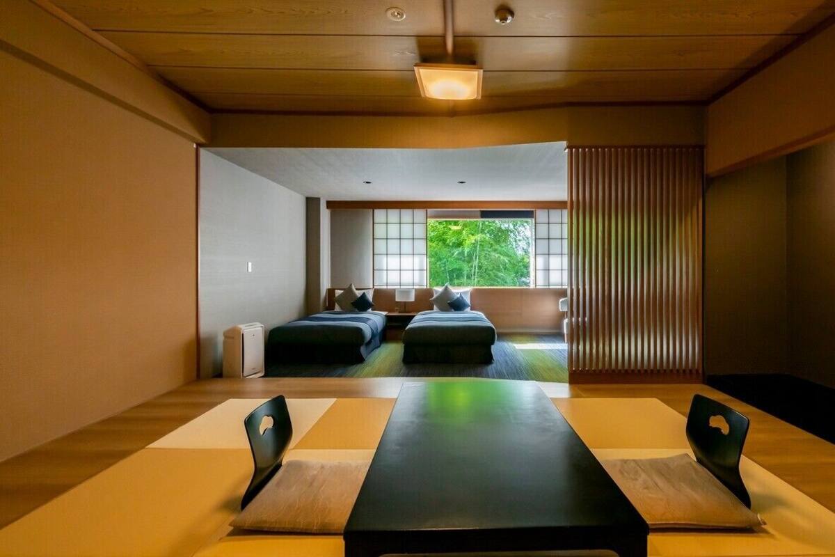 A room with a long table, two beds, and a view of greenery outside.
