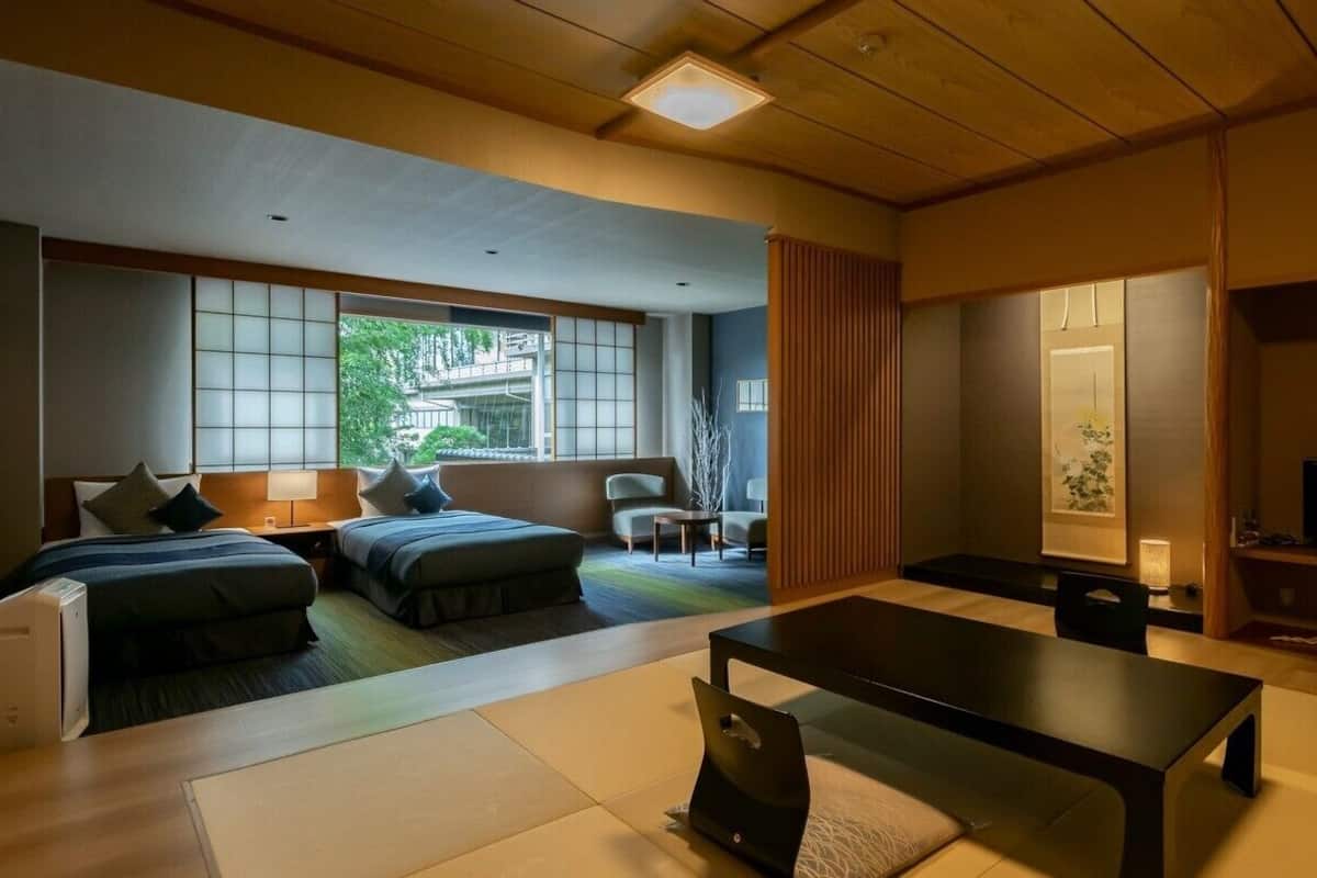 A modern hotel room with a sofa bed, a dining table, a TV, and a view of greenery outside.