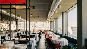 Un restaurant avec des tables et des chaises, un espace cuisine et de grandes fen êtres.