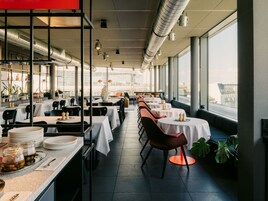 A restaurant with tables and chairs, a kitchen area, and large windows.