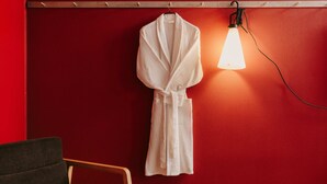 Un mur rouge, une robe blanche suspendue à une tige métallique, une chaise en bois et une lampe sur pied.