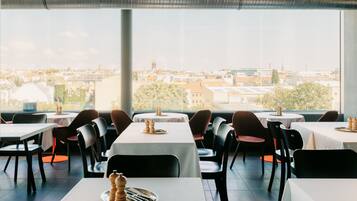 A restaurant with tables and chairs, large windows with a city view, and a ventilation system above.