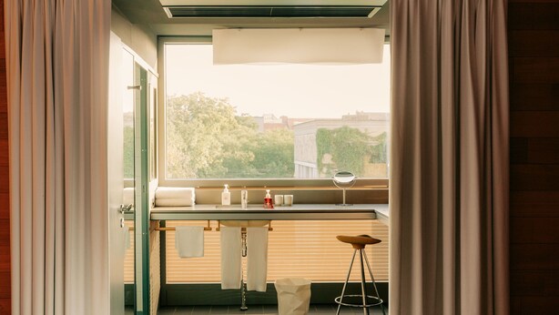 Une chambre d’hôtel avec un lit, une petite table avec des articles de toilette, un tabouret et une vue sur la verdure environnante.