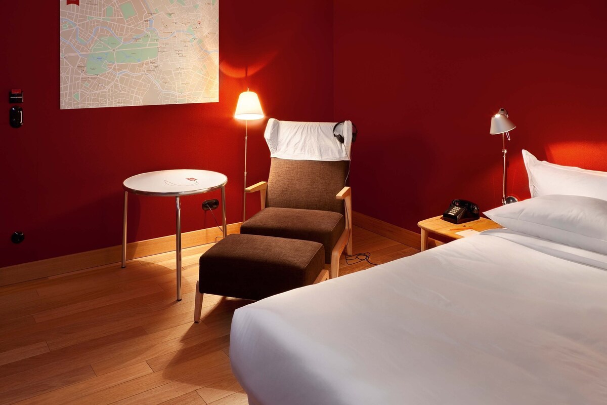 A hotel room with a bed, a chair, a small table, a lamp, and a map on the wall.