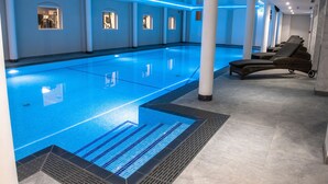 Una piscina interior moderna con agua azul cristalina, sillones de descanso y suelo de baldosas.