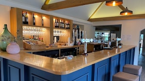 Zona de bar con mostrador de madera, estantes con botellas y vasos, y un área de asientos con taburetes.