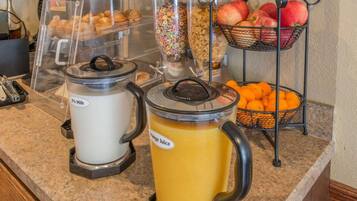Una barra de desayuno con dispensadores de leche y jugo de naranja, una cesta de fruta y un recipiente con cereales.