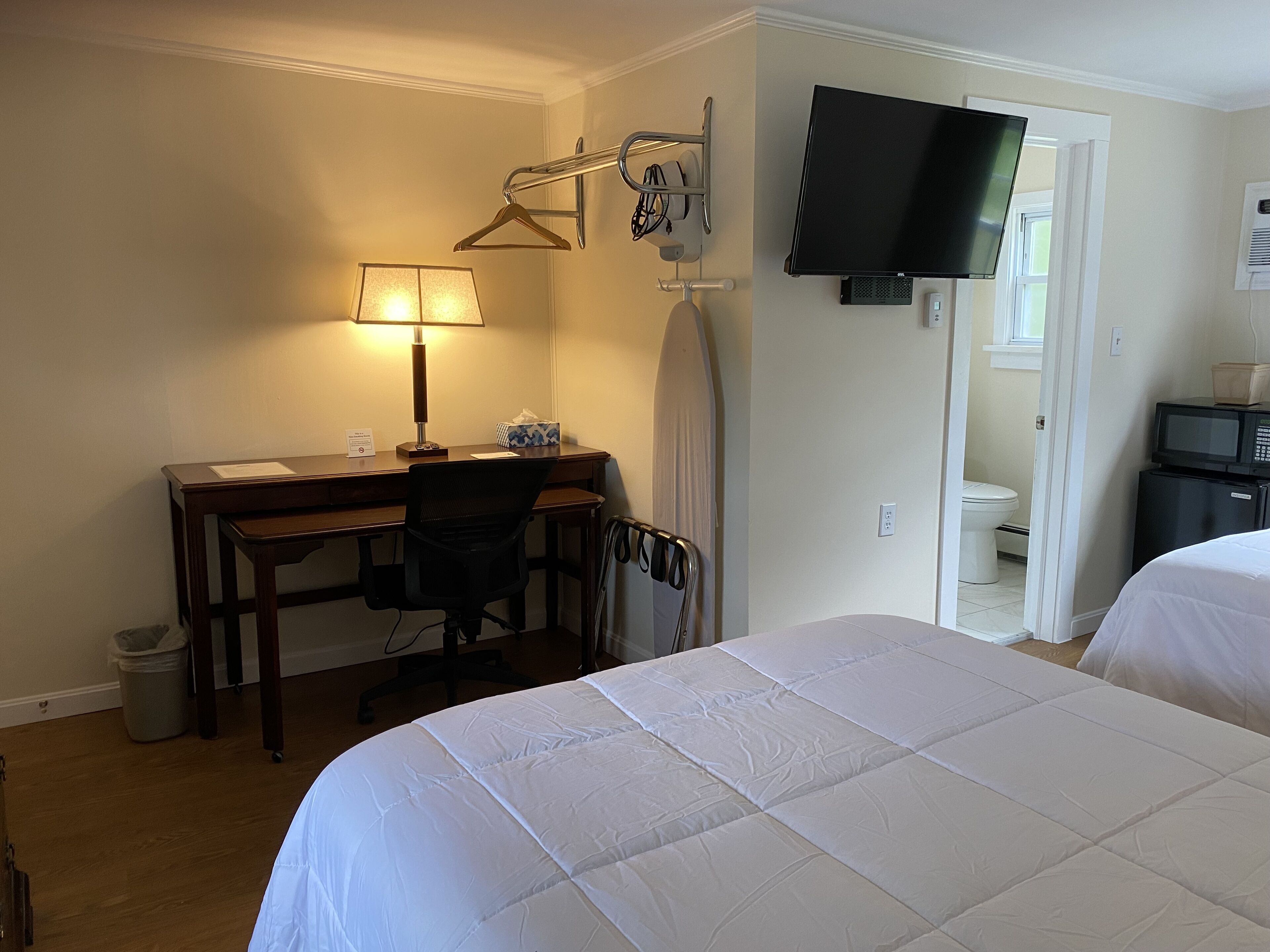 A hotel room with a bed, desk, chair, lamp, TV, and a small kitchenette.