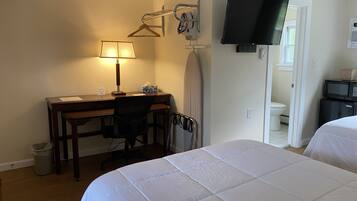 A hotel room with a bed, desk, chair, lamp, TV, and a small kitchenette.