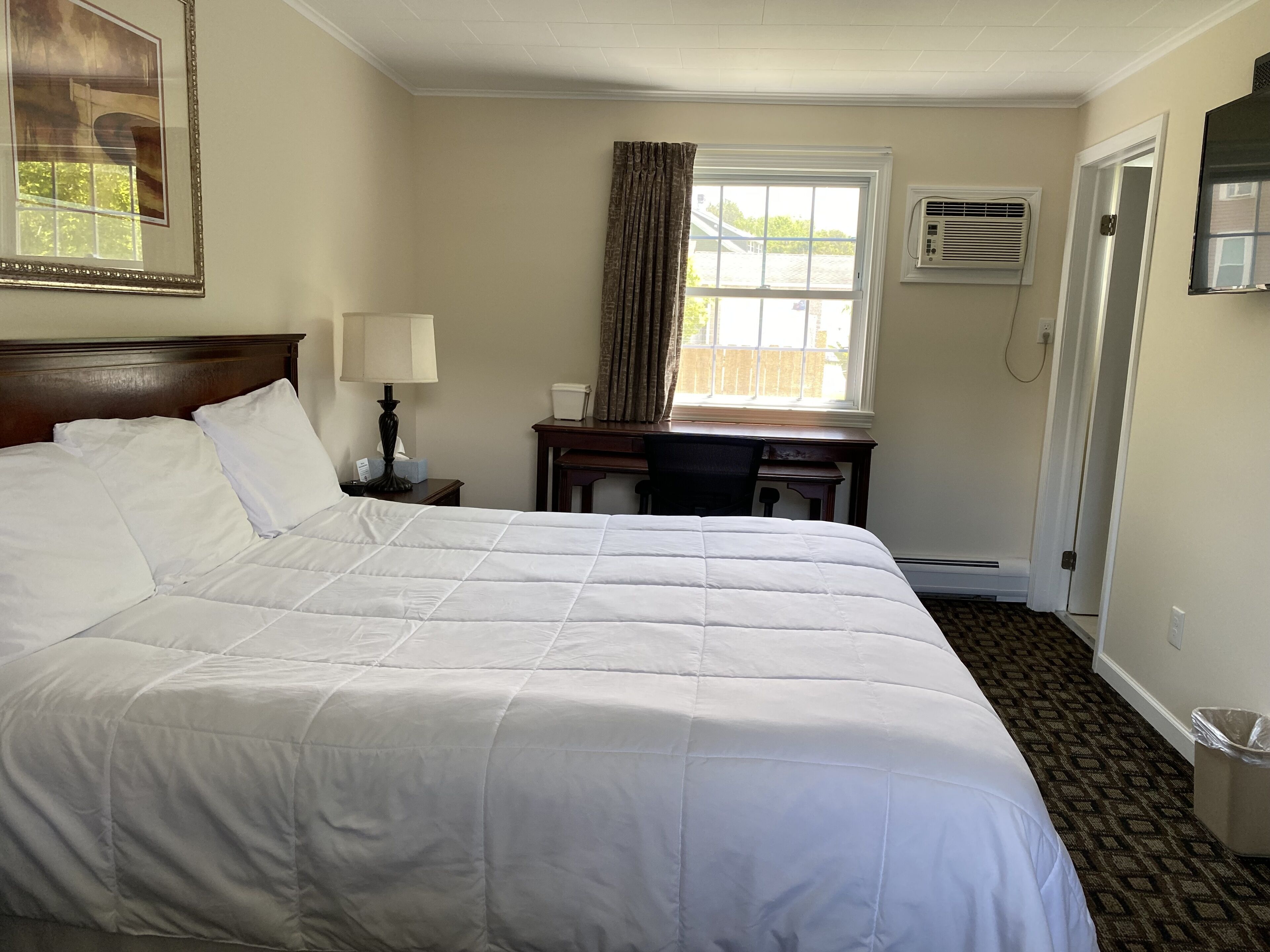 Habitación de hotel con una cama grande, un escritorio, una silla, una lámpara, una ventana con cortinas, un equipo de aire acondicionado y un cuadro en la pared.