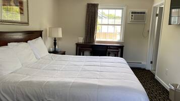 A hotel room with a large bed, a desk, a chair, a lamp, a window with curtains, an air conditioning unit, and a picture on the wall.