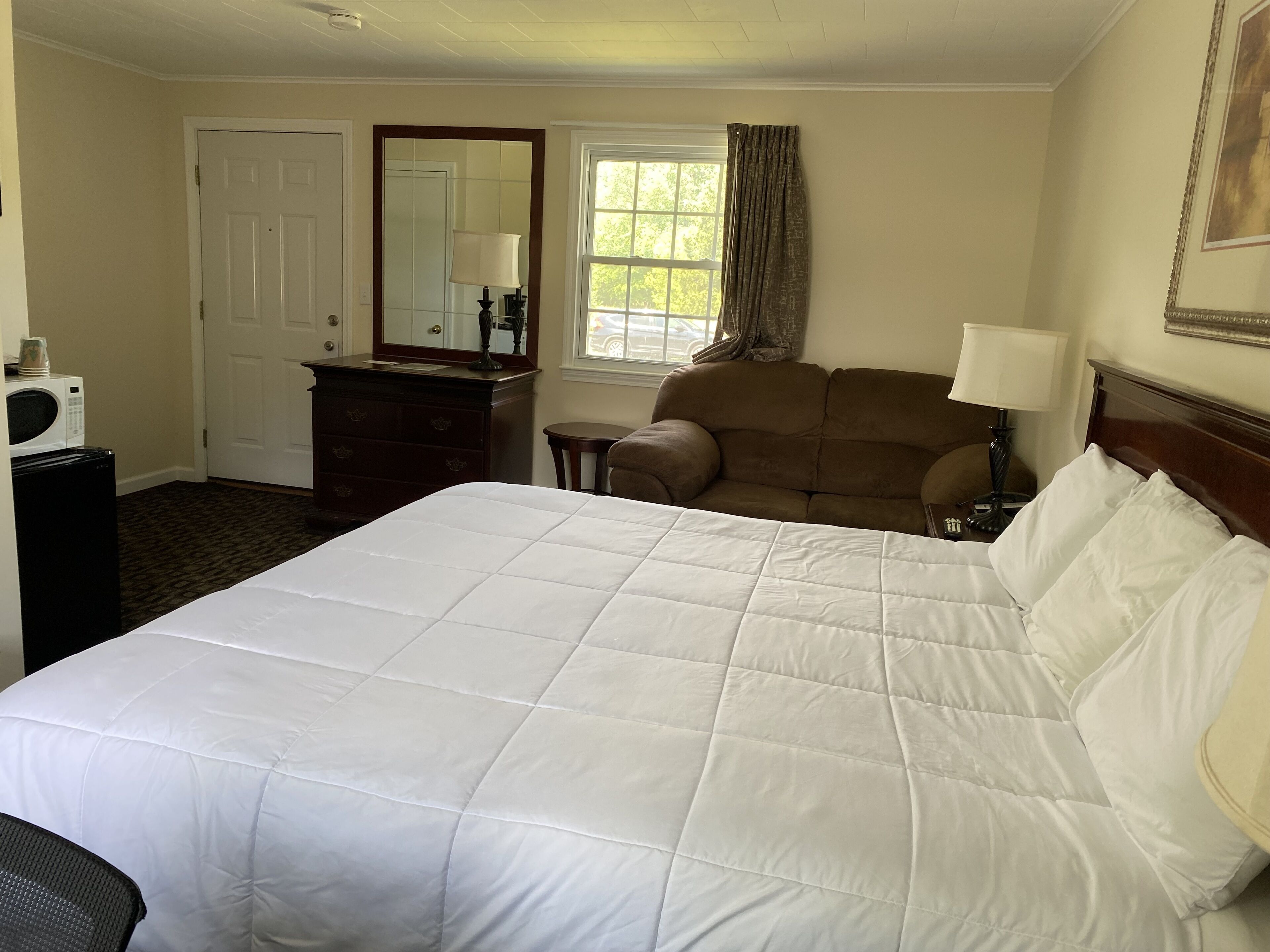 A hotel room with a large bed, a dresser, a chair, a desk with a lamp, and a window with curtains.