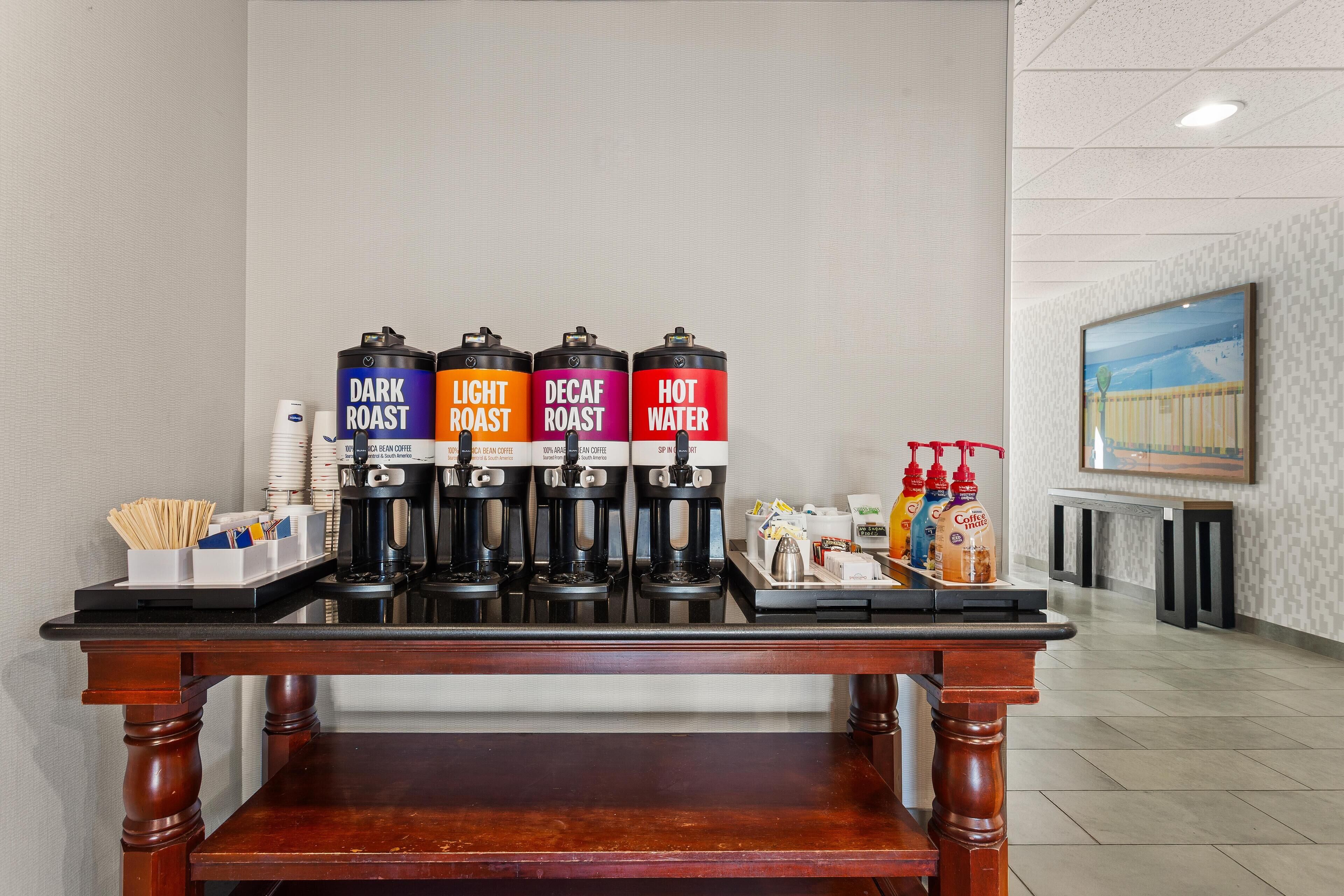 Um espaço de café com quatro dosadores identificados como 'Dark Roast', 'Light Roast', 'Decaf Roast' e 'Água Quente'. Há xícaras de café e uma bandeja com acompanhamentos na mesa.