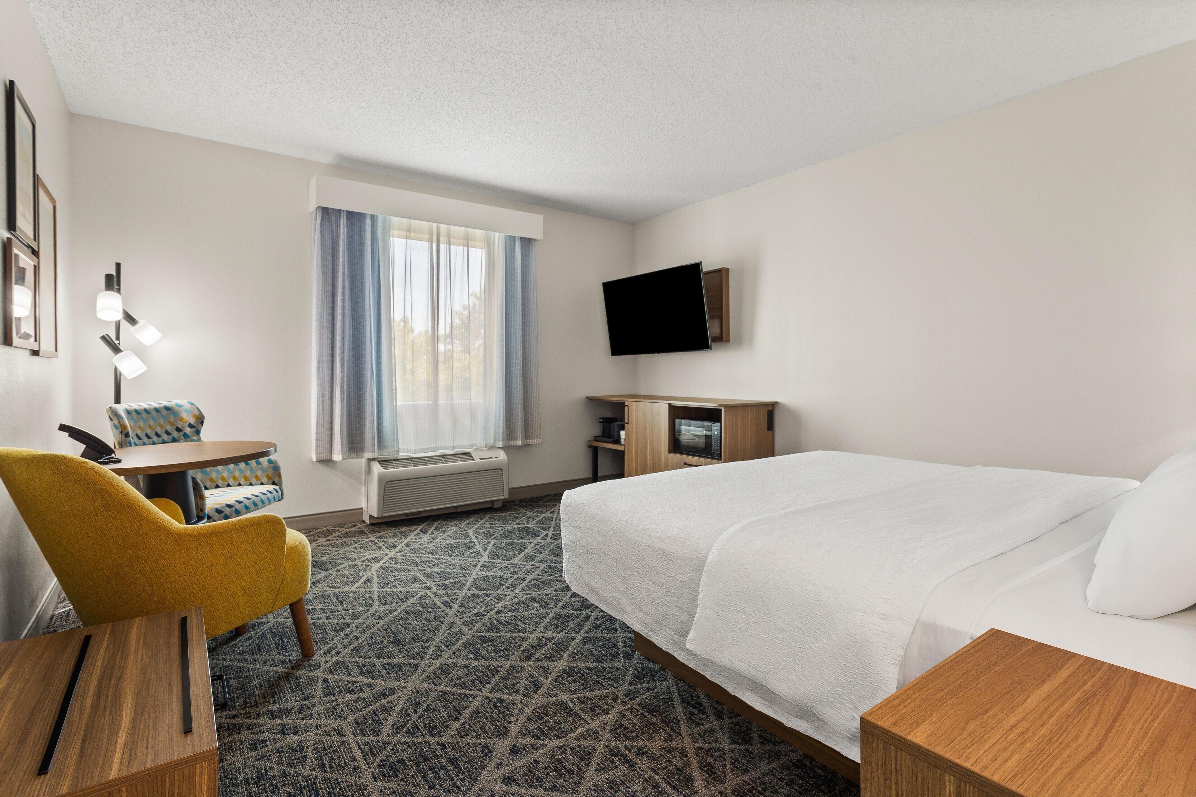 A hotel room with a bed, a desk with a chair, a TV, and a window with curtains.