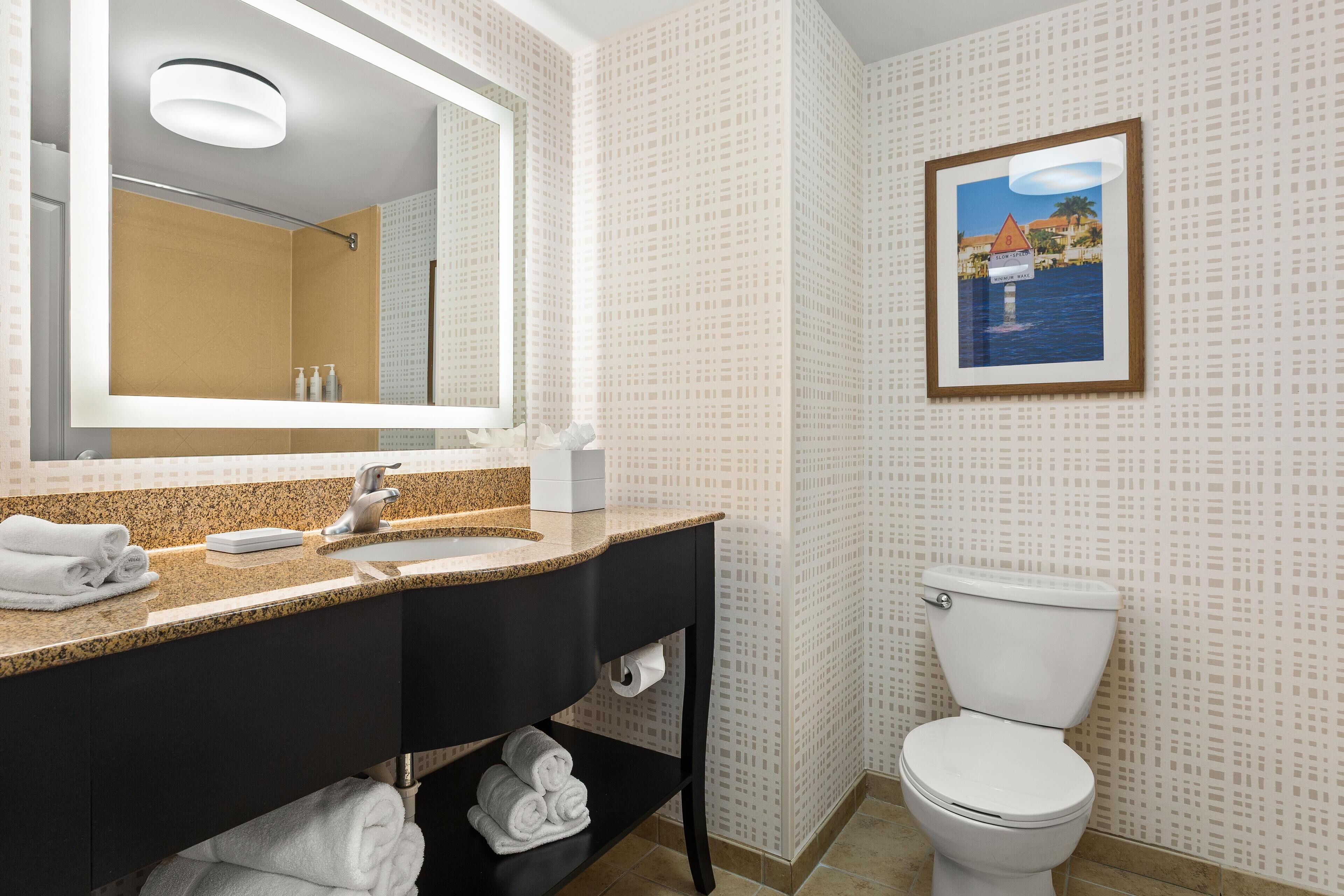 A bathroom with a granite countertop sink, a black vanity, a white toilet, and a framed picture on the wall.