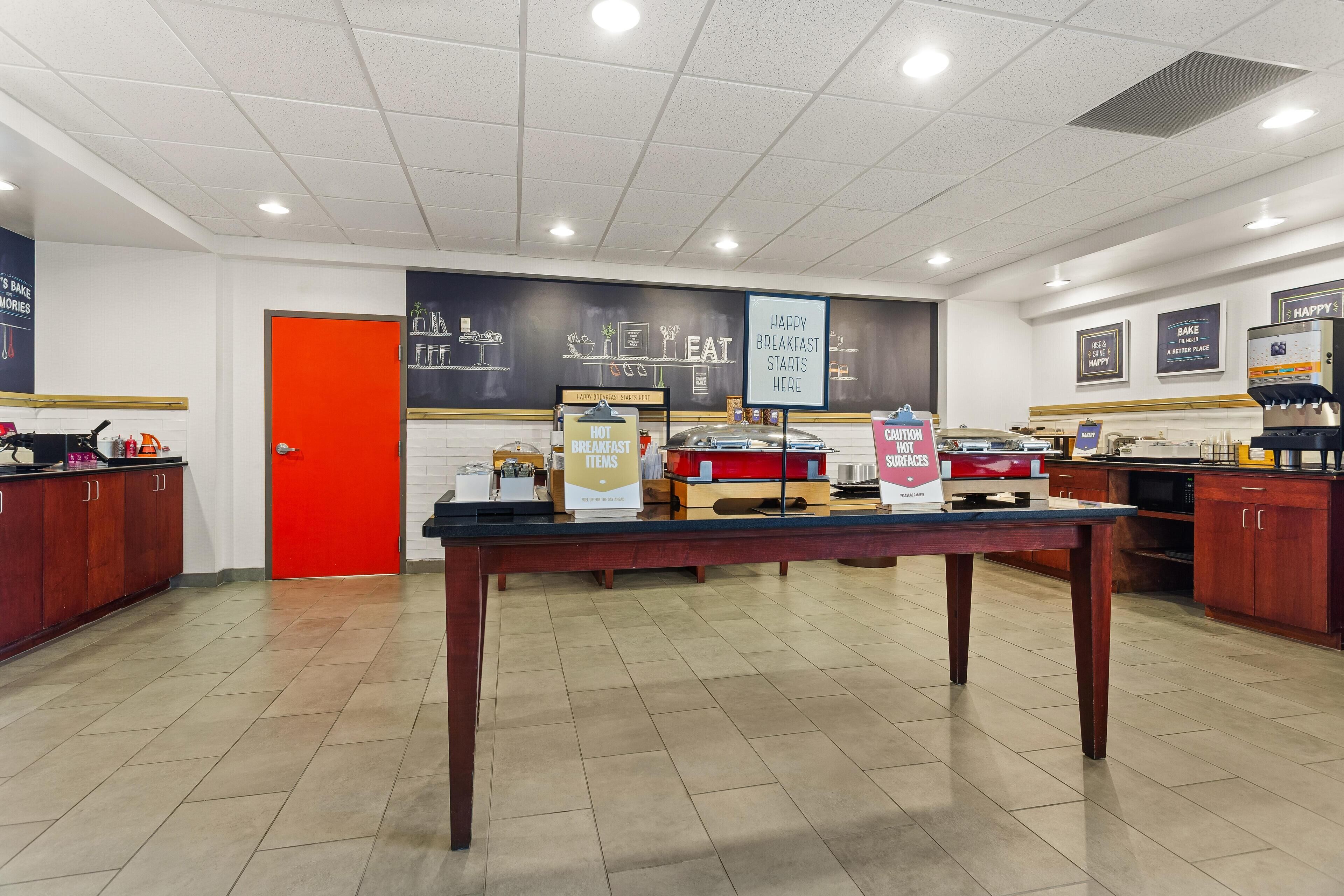 Un espace réservé au petit-déjeuner, avec un comptoir, des panneaux indiquant les plats chauds proposés et précisant qu'il n'est pas nécessaire de réserver, ainsi qu'un mur orné d'une affiche sur laquelle on peut lire : « C'est ici que commence un délicieux petit-déjeuner ».