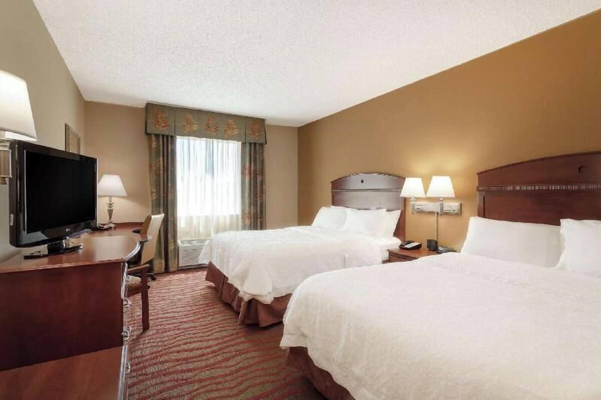 A hotel room with two beds, a desk with a television, a lamp, and a window with curtains.