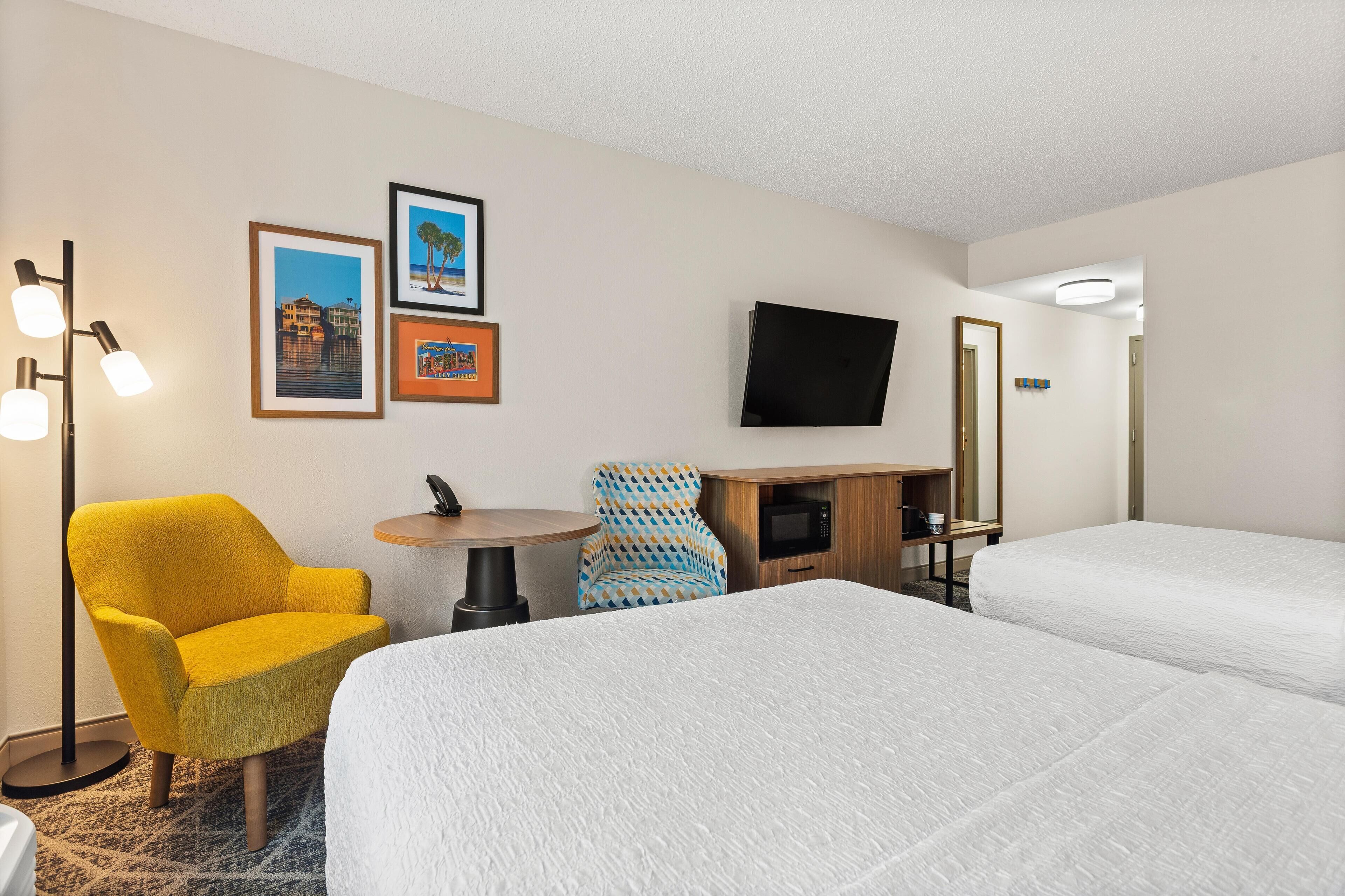 A hotel room with two beds, a yellow armchair, a blue patterned chair, a small round table, and a flat-screen TV mounted on the wall.