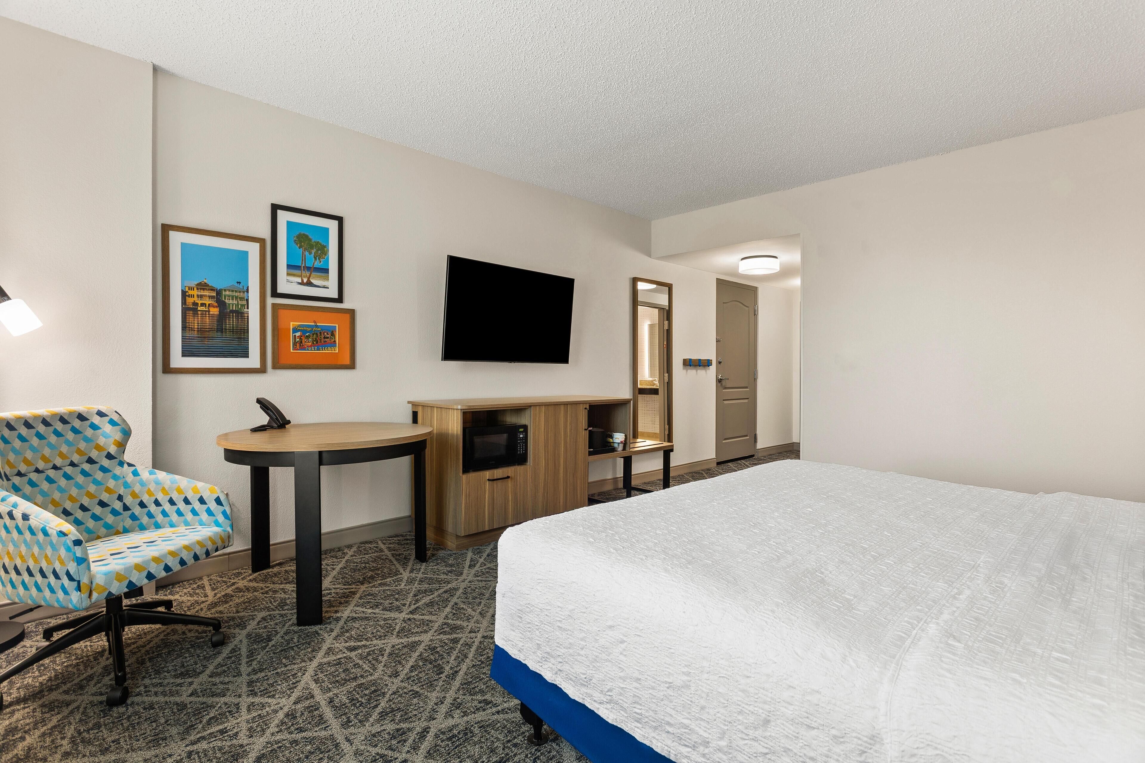 A hotel room with a bed, a desk, a chair, a flat-screen TV, and framed pictures on the wall.