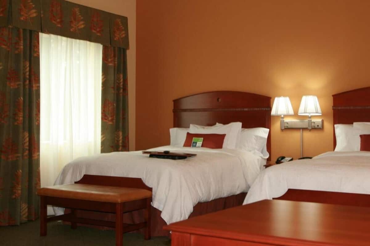 A hotel room with two beds, a bench, a bedside table, and a lamp.