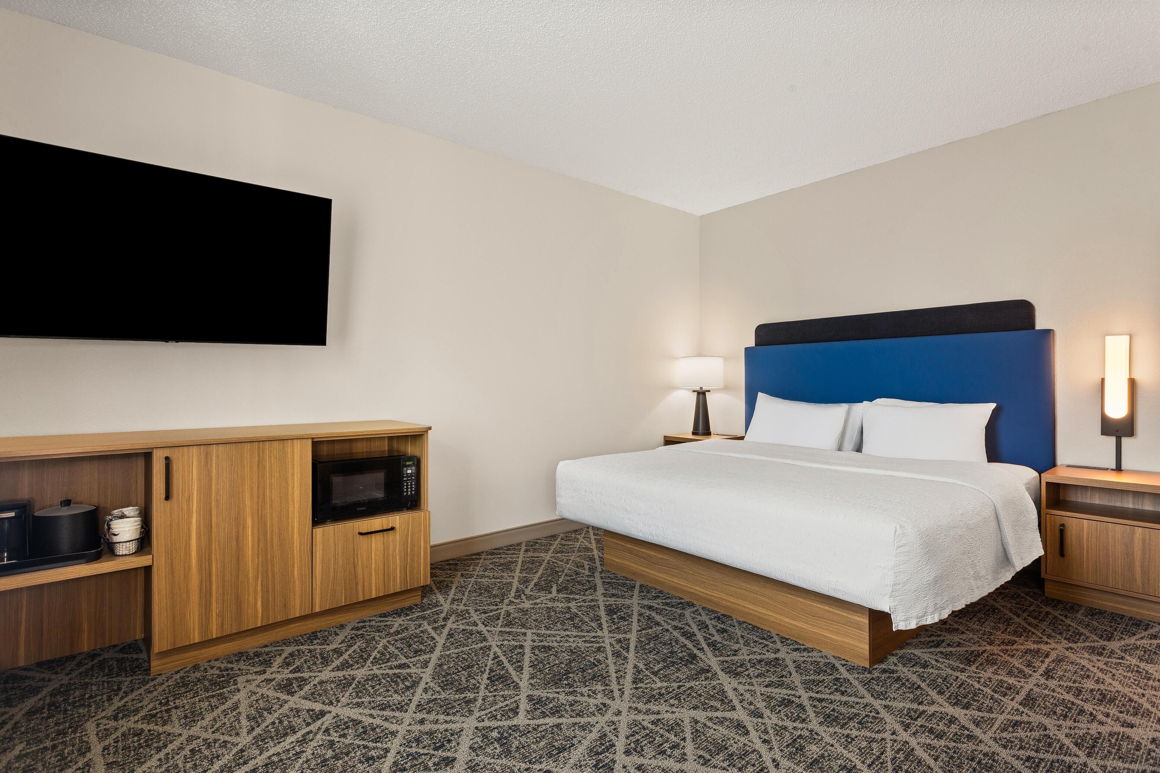 Hotelkamer met een bed, een wandgemonteerde flatscreen-tv, een houten dressoir, een magnetron en een kamerbreed tapijt met dessin.
