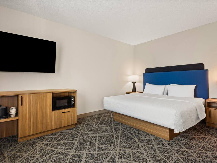 Ein Hotelzimmer mit einem Bett, einem an der Wand befestigten Flachbildfernseher, einer Holzkommode, einer Mikrowelle und einem gemusterten Teppich.