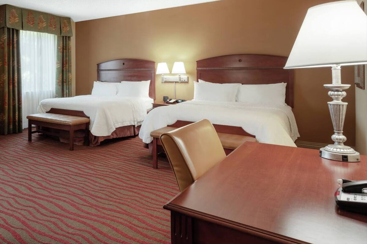 A hotel room with two beds, a desk, and a lamp.