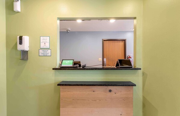 A reception desk with a computer, a check-in/out sign, and a hand sanitizer dispenser. - Extended Stay America Select Suites - Orlando - East (Orlando)