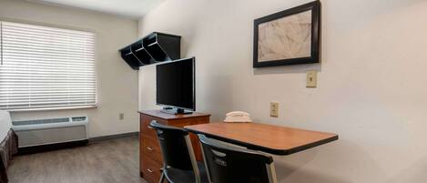 A hotel room with a wooden desk, two black chairs, a flat-screen TV, a telephone, and a framed abstract artwork on the wall.