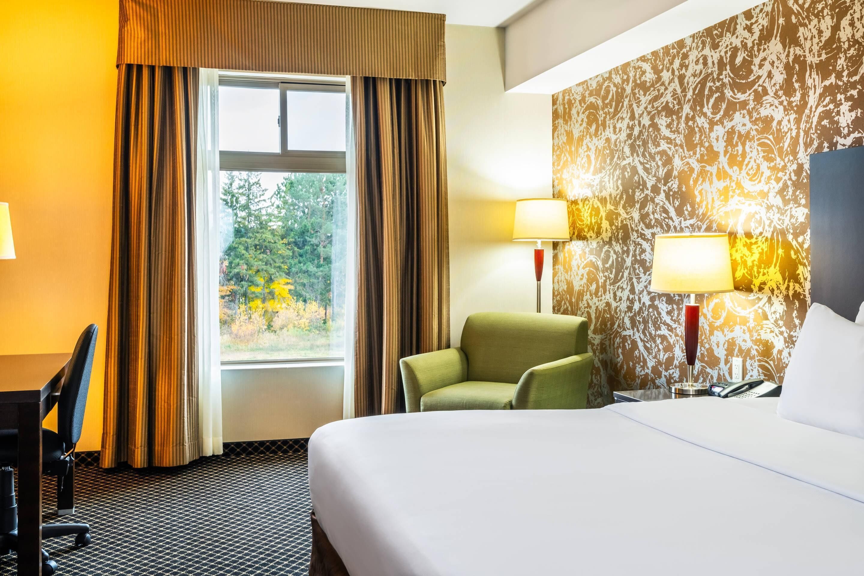 A hotel room with a large bed, a desk with a chair, two bedside lamps, and a view of greenery through the window.