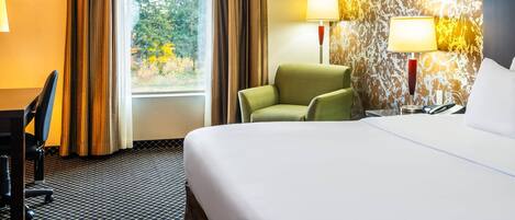 Ein Hotelzimmer mit einem großen Bett, einem Schreibtisch mit Stuhl, zwei Nachttischlampen und Blick ins Grüne durch das Fenster.
