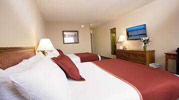 Habitación de hotel con dos camas, cabecero de madera, mesita de noche con lámpara, cómoda, espejo y televisor mostrando un mensaje de bienvenida.