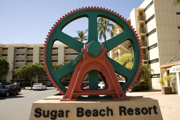 A large gear sculpture with the text "Sugar Beach Resort" in front of a building with multiple balconies and palm trees.