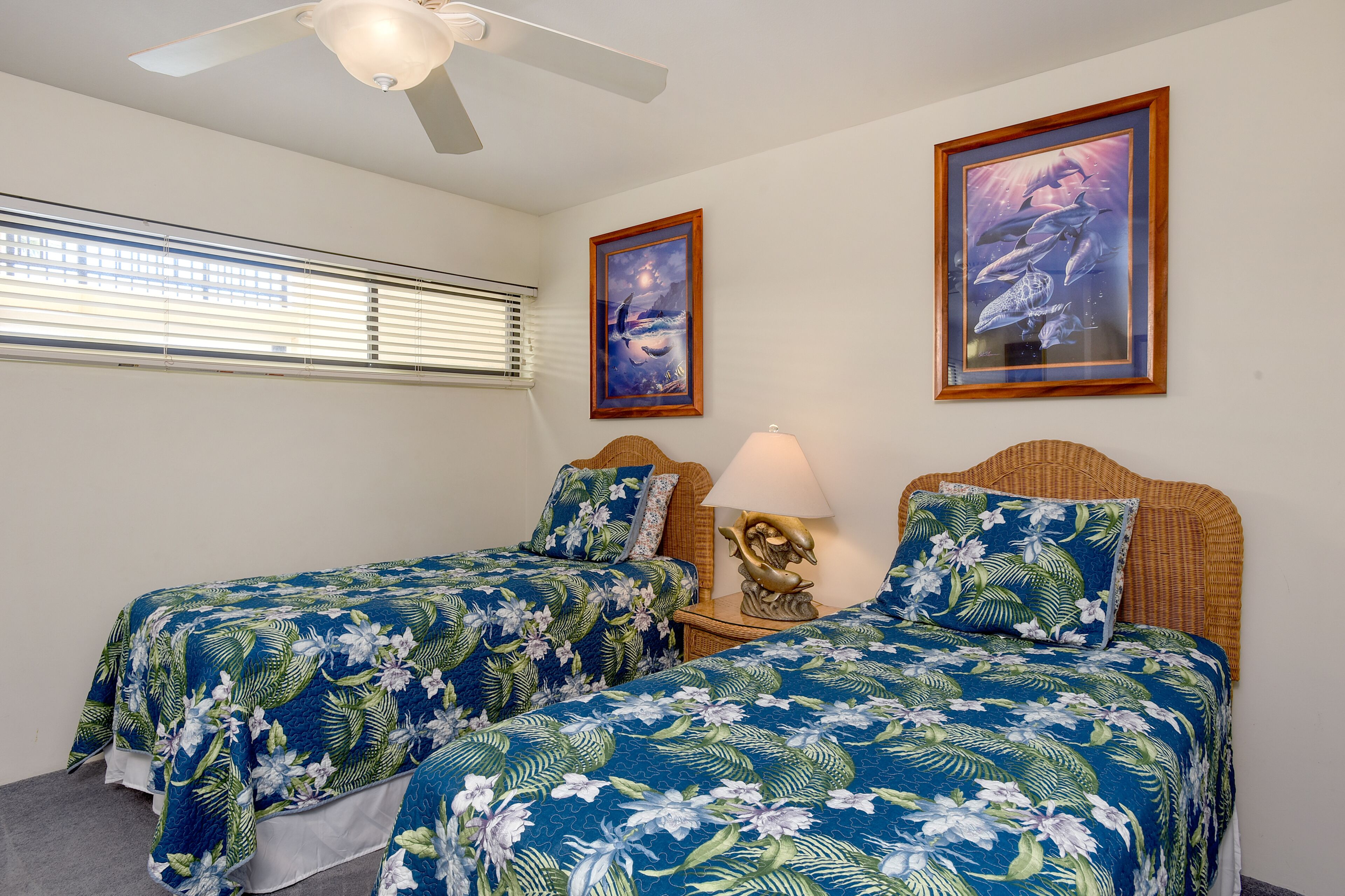 A bedroom with two beds, a ceiling fan, a window with blinds, and two framed pictures on the wall.