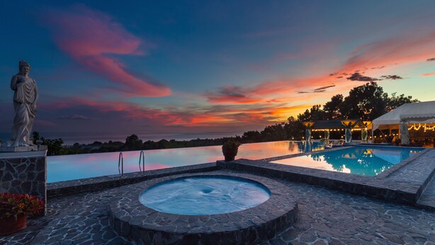 A stone pool with a hot tub, surrounded by a stone deck, overlooking a body of water and a sunset sky. - The Peacock Garden (Baclayon)