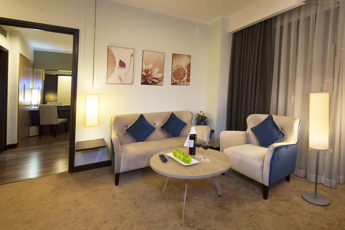 A hotel room with a sofa, two armchairs, a coffee table with apples and wine, and artwork on the wall.