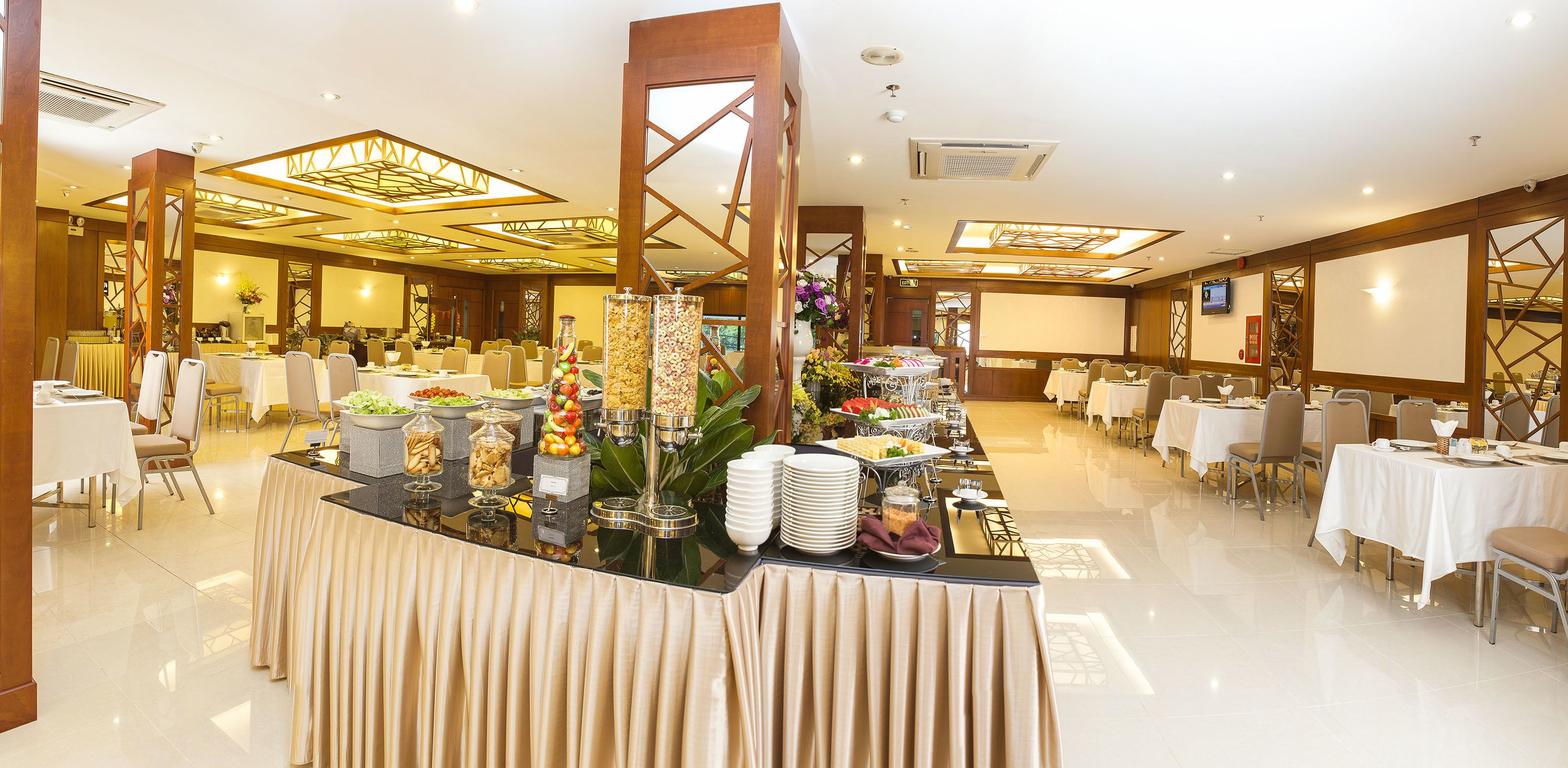 Un amplio salón de banquetes con varias mesas redondas, un bufé central con diversos alimentos y un diseño de techo moderno.