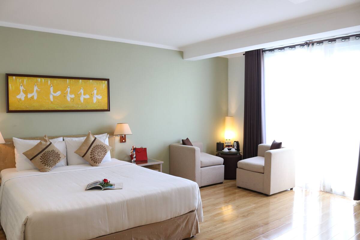 A hotel room with a large bed, a desk, two armchairs, a nightstand, a lamp, and a painting on the wall.